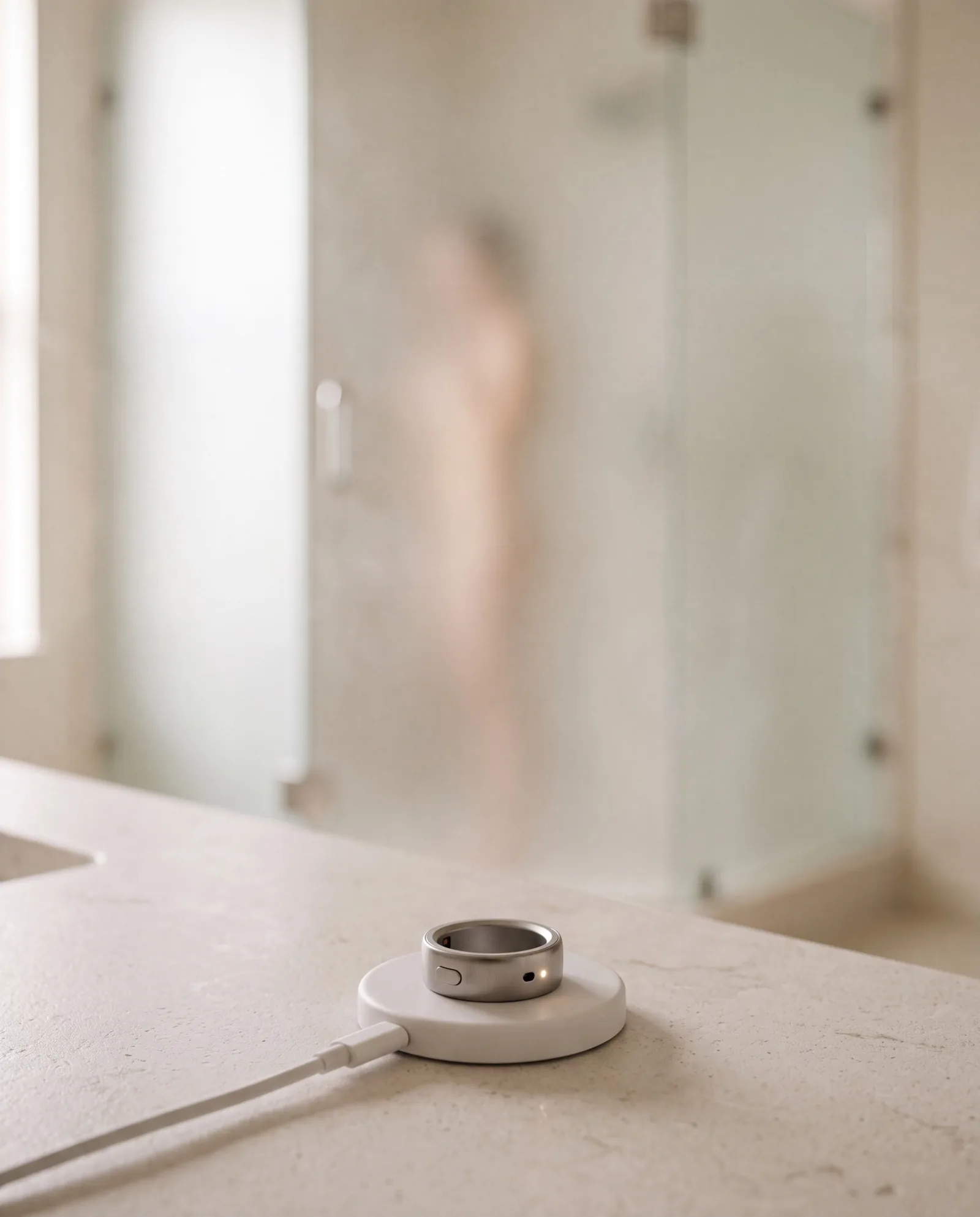 The ring charging on its puck in the foreground, a figure showering behind frosted glass